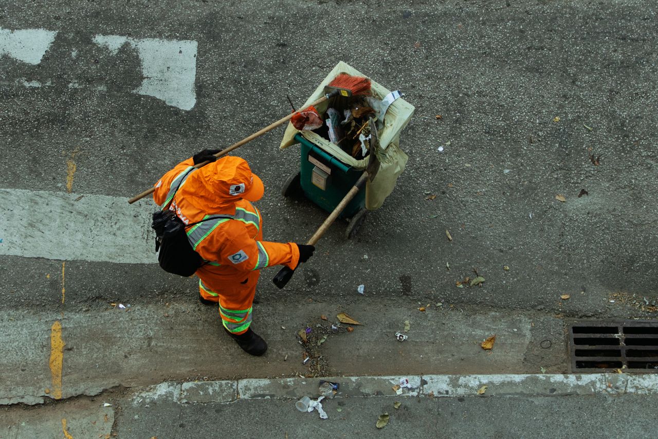 Overhead view of a street cleaner in orange uniform with a cart on a city sidewalk.